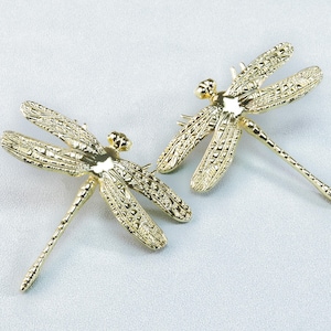 May include: Two gold-colored dragonfly brooches. Each brooch features detailed wing patterns and a slender body. The brooches are displayed on a light-colored surface, showcasing their intricate design and metallic sheen.