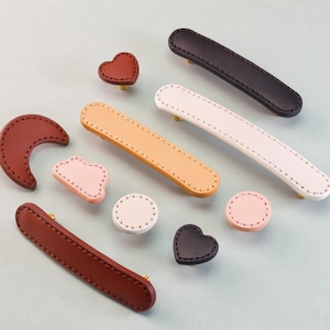 May include: A collection of leather cabinet pulls and knobs in various shapes and colors. Includes brown, black, white, and tan pulls, along with heart, moon, and round knobs. Each piece features decorative stitching and gold-toned hardware.