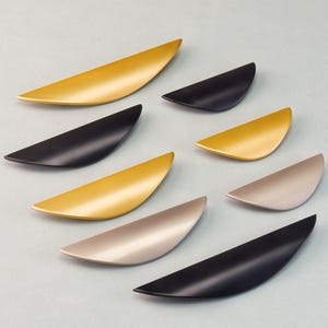May include: An assortment of cabinet pulls in various colors, including gold, black, and silver. The pulls have a curved, crescent shape and are arranged on a light blue surface. These hardware pieces are designed for furniture.