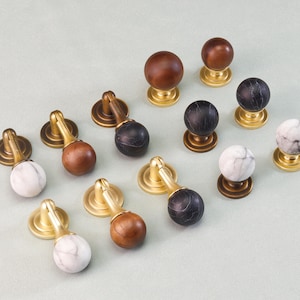 May include: A collection of cabinet knobs with brass bases and various tops. The tops include wood, marble, and black marble. The knobs are arranged on a light green surface, showcasing their different styles and finishes.
