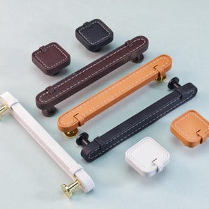 May include: A collection of leather cabinet hardware in various colors, including white, brown, tan, and black. The hardware includes square knobs and rectangular pulls with white stitching and gold or black hardware.