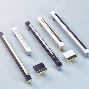 May include: A selection of cabinet handles and knobs in various designs. The pulls are rectangular, and the knobs are square. The colour schemes include black, white, silver, and gold. The hardware is displayed on a light blue surface.