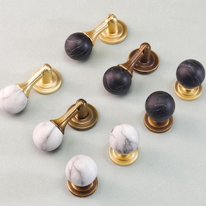 May include: A collection of cabinet hardware, including knobs and pulls. The knobs and pulls feature marble-like spheres in white and black, paired with gold and bronze-toned metal bases and handles. The hardware is arranged on a light gray surface.