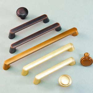 Op de afbeelding: Een assortiment kastbeslag in diverse metallic afwerkingen. De selectie omvat handgrepen en knoppen in brons, goud en messing. Het beslag is gerangschikt op een lichtblauwe ondergrond.