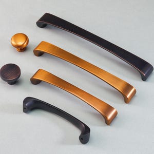 Op de afbeelding: Een collectie van kastbeslag in diverse metallic afwerkingen. De collectie bevat twee knoppen en drie handgrepen in brons, koper en zwart. Het beslag wordt getoond op een lichtgrijze ondergrond, waarbij de verschillende stijlen en kleuren worden getoond.