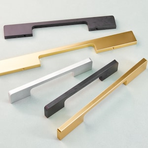 Pode incluir: Uma variedade de puxadores de armário modernos em vários acabamentos metálicos. A seleção inclui preto fosco, dourado, prateado e bronze escovado. Cada puxador é retangular com uma alça embutida, mostrando um design minimalista.