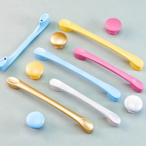 May include: A selection of colourful cabinet hardware, including blue, yellow, pink, gold, and white handles and knobs. The handles are approximately 15 cm long, and the knobs are round. The hardware is arranged on a light grey surface.