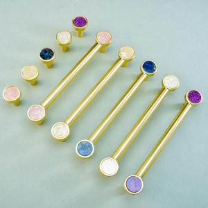 Könnte beinhalten: Eine Sammlung von Schrankbeschlägen, darunter goldfarbene Stangengriffe und passende Knöpfe. Die Griffe und Knöpfe haben farbenfrohe, runde Akzente in Rosa, Weiß, Blau und Lila. Die Griffe sind ca. 15 cm lang.