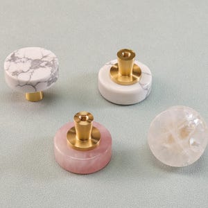 May include: Four decorative cabinet knobs. One is white with gray veining, another is white with a brass base, a pink knob with a brass base, and a clear, spherical knob. These knobs are for home decor.