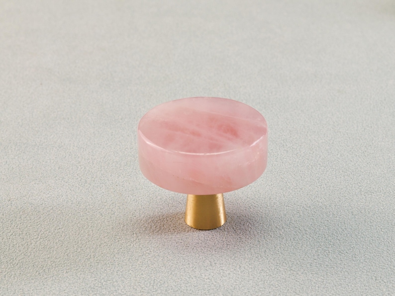 May include: A rose quartz cabinet knob with a gold-colored base. The knob is a light pink color and has a smooth, rounded top. The base is cylindrical and made of a shiny, gold-toned metal. The knob is designed for use on cabinets or drawers.