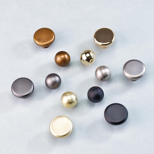 May include: A selection of cabinet knobs in diverse metallic finishes, including gold, bronze, silver, and black. The knobs are round and have varied textures and shapes, suitable for furniture hardware.