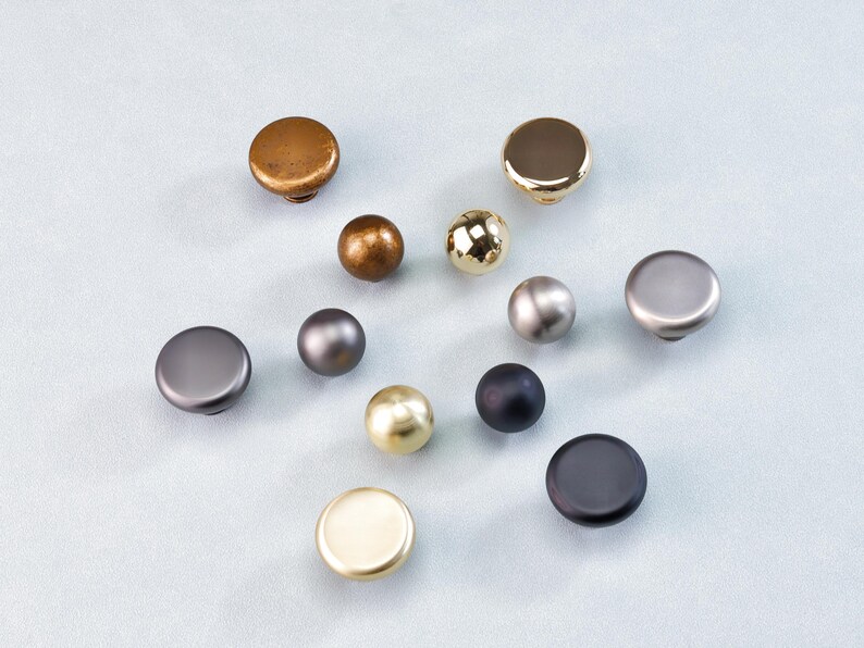 May include: A selection of cabinet knobs in diverse metallic finishes, including gold, bronze, silver, and black. The knobs are round and have varied textures and shapes, suitable for furniture hardware.