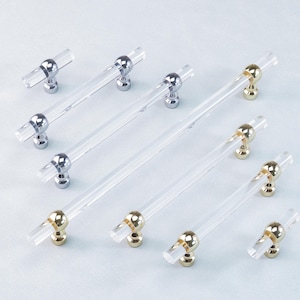 May include: Clear acrylic cabinet pulls with silver and gold-toned hardware. The pulls have a cylindrical design with a clear, transparent body and a T-shaped handle. The hardware is either silver or gold, adding a touch of elegance to the design.