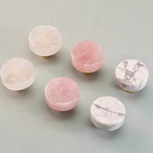 May include: Six decorative cabinet knobs in various colors and patterns. The knobs are round with brass stems. Colors include white, pink, and white with gray veining. These knobs are made of natural stone.