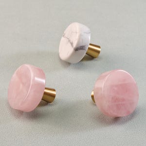 May include: Three decorative cabinet knobs. Two are pink, and one is white with gray veining. Each knob has a round, polished stone top and a brass-colored stem.