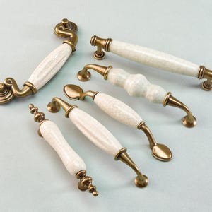 Może przedstawiać: Asortyment uchwytów do szafek w stylu antycznym z białymi ceramicznymi uchwytami i mosiężnymi metalowymi okuciami. Uchwyty różnią się kształtem i wzornictwem, w tym zakrzywione, cylindryczne i dekoracyjne końcówki. Ceramiczne uchwyty mają spękaną glazurę.