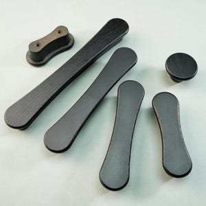 May include: A collection of black wooden cabinet hardware, including handles and a knob. The handles vary in size and shape, with rounded ends and a slightly curved design. The hardware is set against a light background.