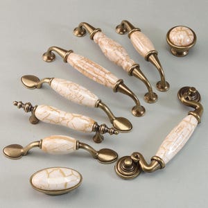 Könnte beinhalten: Eine Sammlung von Schrankbeschlägen, darunter Griffe und ein Knauf. Die Griffe haben eine cremefarbene, marmorähnliche Oberfläche mit goldfarbenen Metallakzenten. Geeignet für Möbel- und Heimdekorationsprojekte.
