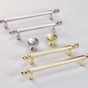 May include: An assortment of cabinet hardware, including silver and gold-toned handles and knobs. The handles have a ribbed design and decorative ends. The knobs have a fluted design. The hardware is displayed on a light gray surface.