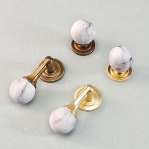 May include: Four cabinet knobs with white and gray marble spheres and brass or gold-toned bases. Two knobs have a curved design, while the other two are spherical. The knobs are arranged on a light gray surface.