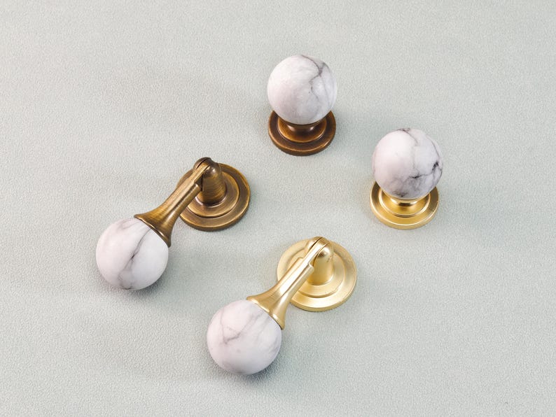 May include: Four cabinet knobs with white and gray marble spheres and brass or gold-toned bases. Two knobs have a curved design, while the other two are spherical. The knobs are arranged on a light gray surface.
