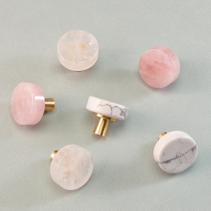 May include: A collection of decorative cabinet knobs in various colors and materials. The knobs feature rose quartz, white marble with gray veining, and translucent stone. Each knob has a gold-colored metal base.