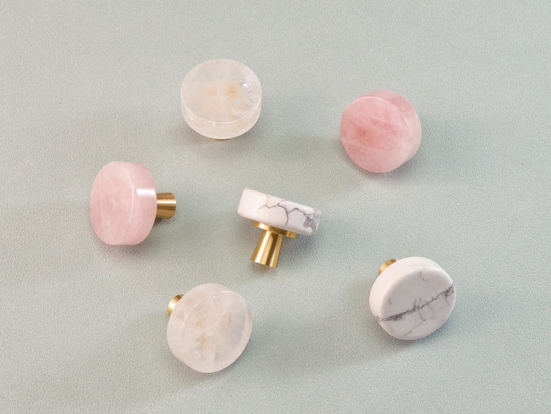 May include: A collection of decorative cabinet knobs in various colors and materials. The knobs feature rose quartz, white marble with gray veining, and translucent stone. Each knob has a gold-colored metal base.