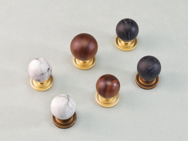 May include: Six decorative cabinet knobs with a round shape. The knobs feature a variety of materials, including wood and marble, and are mounted on brass or bronze bases. The knobs are in shades of brown, black, and white.