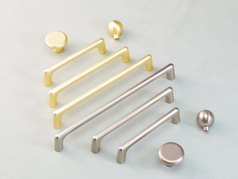 May include: A selection of cabinet hardware, featuring gold and silver-toned handles and knobs. The handles are rectangular with curved ends, and the knobs are round or spherical. The hardware is displayed on a light blue surface.