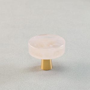 May include: A cabinet knob with a round, light pink top and a gold-colored base. The knob has a smooth, polished finish and is designed for furniture. The hardware is a decorative accent for drawers and cabinets.