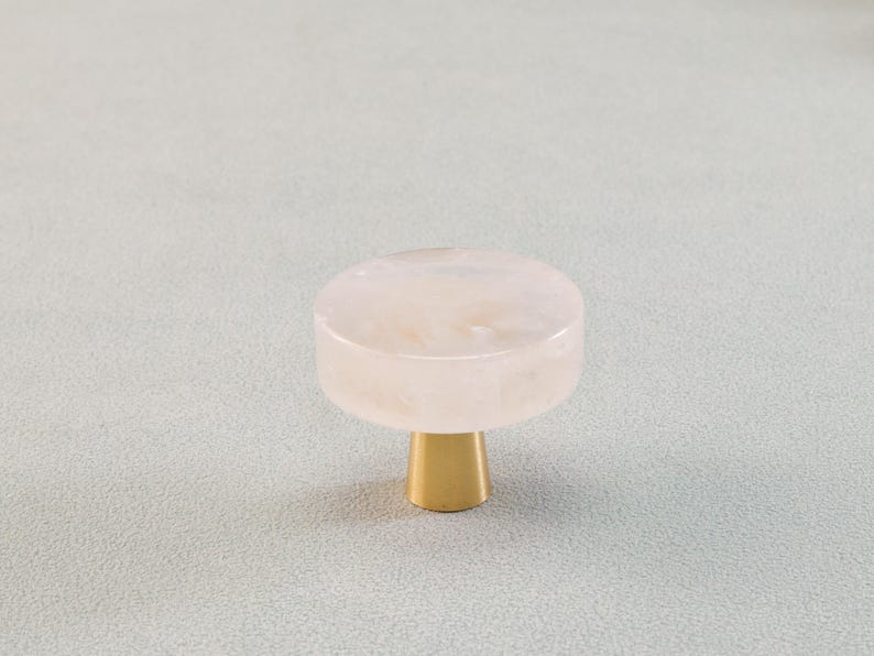 May include: A cabinet knob with a round, light pink top and a gold-colored base. The knob has a smooth, polished finish and is designed for furniture. The hardware is a decorative accent for drawers and cabinets.