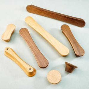 May include: An assortment of wooden cabinet hardware in various shapes and shades. The collection includes knobs, pulls, and handles in light, medium, and dark wood tones. The hardware is arranged on a light blue surface, showcasing the natural wood grain.