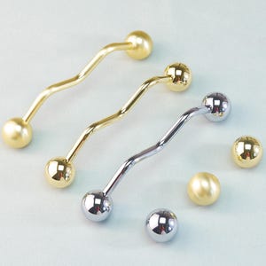 May include: Three wavy metal drawer pulls in gold and silver, each with a ball on either end. Two additional gold and silver balls are also present. The hardware is likely for cabinets or furniture.