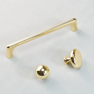 May include: A collection of three gold-coloured cabinet fittings. The set comprises a long, straight handle, a spherical knob, and an oval-shaped knob. The hardware is made of a shiny, reflective metal.