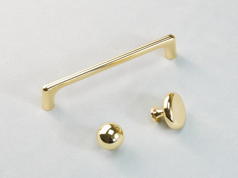 May include: A collection of three gold-coloured cabinet fittings. The set comprises a long, straight handle, a spherical knob, and an oval-shaped knob. The hardware is made of a shiny, reflective metal.