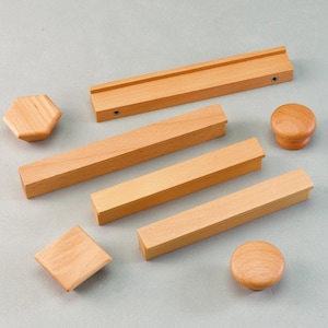 May include: A selection of unfinished wooden cabinet hardware, including knobs and pulls. The hardware features various shapes: hexagonal, square, and round knobs, and rectangular pulls. The wood has a light, natural colour.