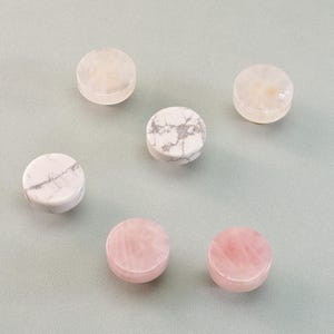 May include: Six round, polished gemstones in shades of pink, white, and cream are arranged on a light gray surface. The stones vary in color and veining, with some displaying a marbled effect.