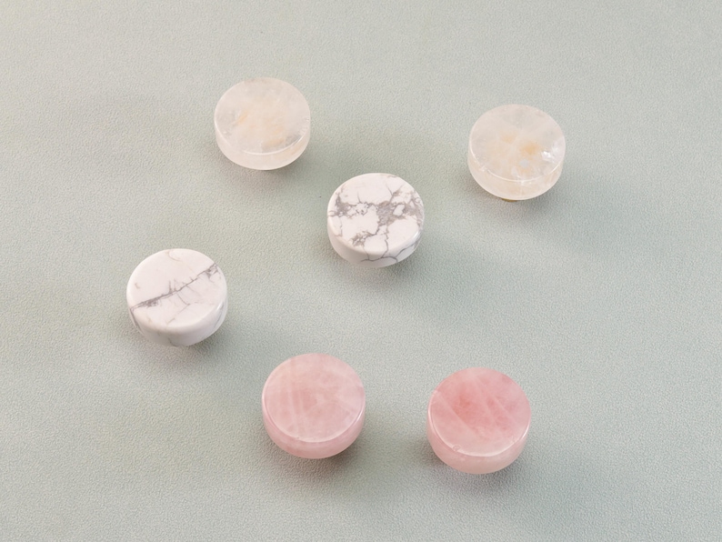 May include: Six round, polished gemstones in shades of pink, white, and cream are arranged on a light gray surface. The stones vary in color and veining, with some displaying a marbled effect.