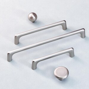 May include: A selection of brushed nickel cabinet hardware, comprising two round knobs and three bar pulls of different lengths. The hardware is presented on a pale blue surface, highlighting a contemporary design style.