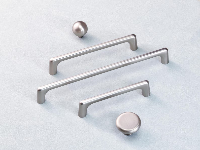 May include: A selection of brushed nickel cabinet hardware, comprising two round knobs and three bar pulls of different lengths. The hardware is presented on a pale blue surface, highlighting a contemporary design style.