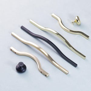 May include: A selection of cabinet handles and knobs in gold and black. The image shows several wavy, elongated pulls in gold and black, along with a gold knob and a black knob. The hardware is displayed on a light grey surface.
