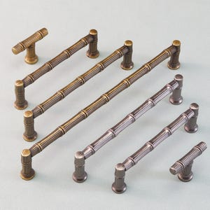 Op de afbeelding: Een collectie bamboe-stijl kastgrepen en knoppen in messing en pewter afwerkingen. De afbeelding toont zowel stanggrepen als knoppen, geschikt voor keuken- of badkamerkasten. De hardware is van metaal.