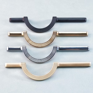 Op de afbeelding: Vier kastgrepen in verschillende metallic afwerkingen: zwart, goud, zilver en messing. Elke greep heeft een gebogen, gegroefde handgreep en rechthoekige uiteinden. De hardware is ontworpen voor meubels en woondecoratie.