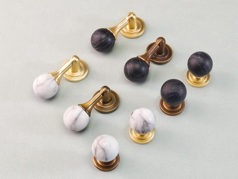 May include: A collection of cabinet hardware, including knobs and pulls. The knobs and pulls feature marble-like spheres in white and black, paired with gold and bronze-toned metal bases and handles. The hardware is arranged on a light gray surface.