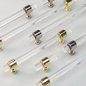 Puede incluir: Tiradores de armario de acrílico transparente con detalles metálicos dorados y plateados. Los tiradores son cilíndricos con un cuerpo transparente y extremos metálicos. Herrajes para muebles y armarios.