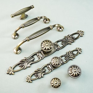 May include: An assortment of antique bronze cabinet hardware, including handles and knobs. The handles have a curved design with ribbed detailing, while the knobs feature a floral center. The hardware is displayed on a light blue background.
