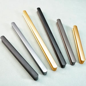 May include: An assortment of modern cabinet pulls in various metallic finishes, including black, silver, gold, and bronze. The pulls are rectangular with rounded ends, arranged diagonally on a light blue surface.