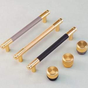 Może przedstawiać: Kolekcja złotych okuć do szafek, w tym trzy uchwyty prętowe z teksturowanymi uchwytami w kolorze szarym, złotym i czarnym oraz trzy pasujące okrągłe gałki. Okucia są prezentowane na jasnoniebieskiej powierzchni.