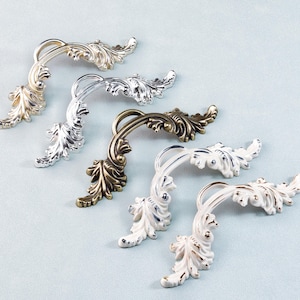 May include: Decorative cabinet pulls in various metallic finishes, including gold, silver, bronze, and white. Each pull features a curved handle with an ornate leaf design, adding a touch of elegance to furniture.