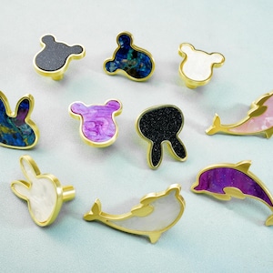 May include: A collection of decorative cabinet knobs in various shapes, including bunnies, dolphins, and bear-like figures. The knobs feature gold-colored metal bases and colorful, patterned inlays in shades of blue, purple, black, and white.
